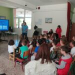 Φωτογραφία στο προαύλιο του σχολείου που απεικονίζει προσωπικό της Μονάδας που κάνει δραστηριότητες με τα παιδιά και συγκεκριμένα κουίζ κάνοντας χρήση διαδραστικής οθόνης